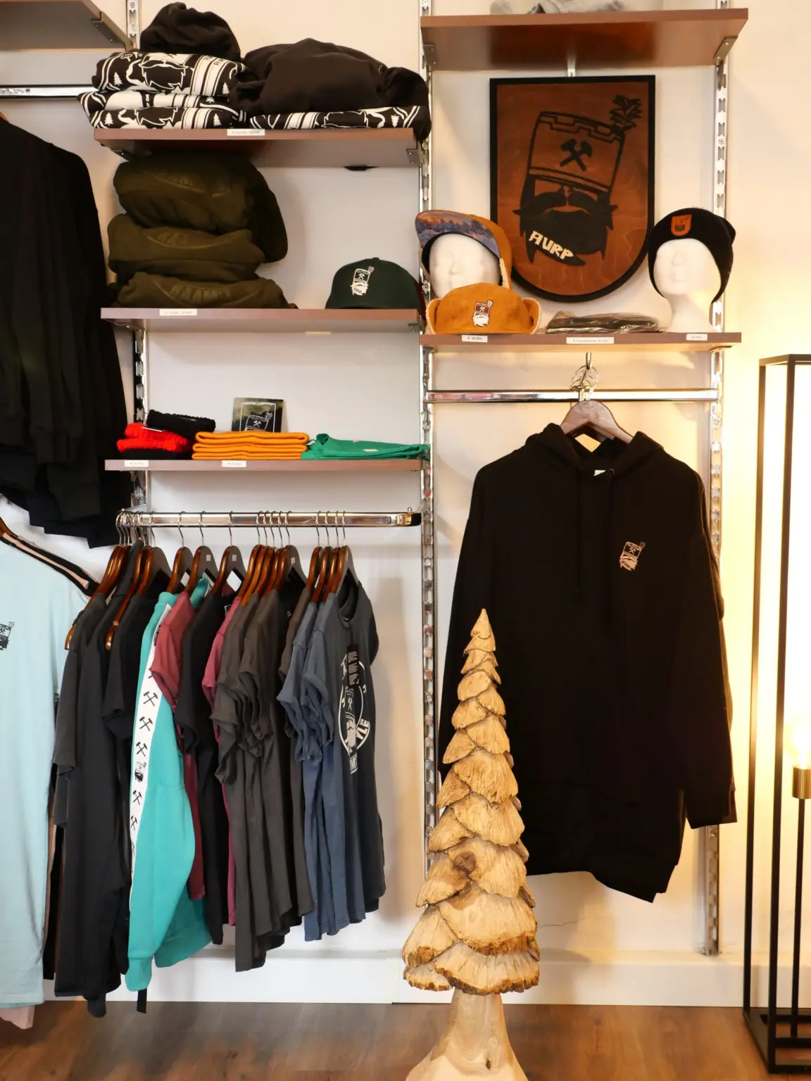 Kleidung im Laden am Markt: Hoodies, T-Shirts und Mützen, teils mit Bergbau-Motiv Schlägel und Eisen, daneben eine geschnitzte Holztanne