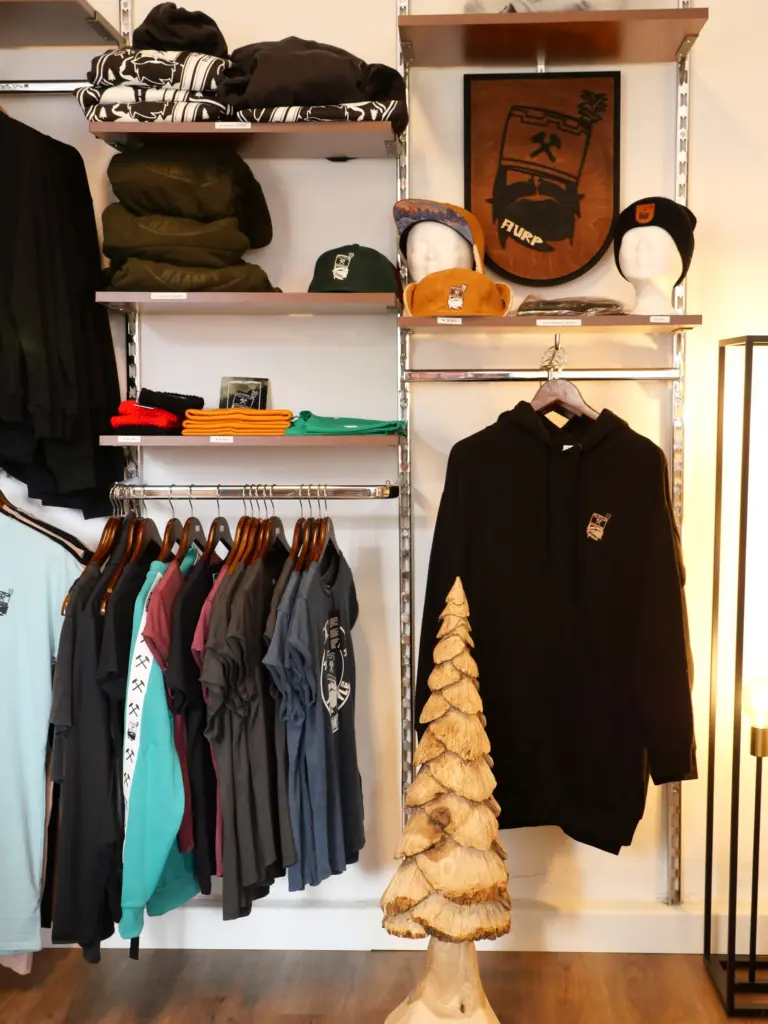 Kleidung im Laden am Markt: Hoodies, T-Shirts und Mützen, teils mit Bergbau-Motiv Schlägel und Eisen, daneben eine geschnitzte Holztanne