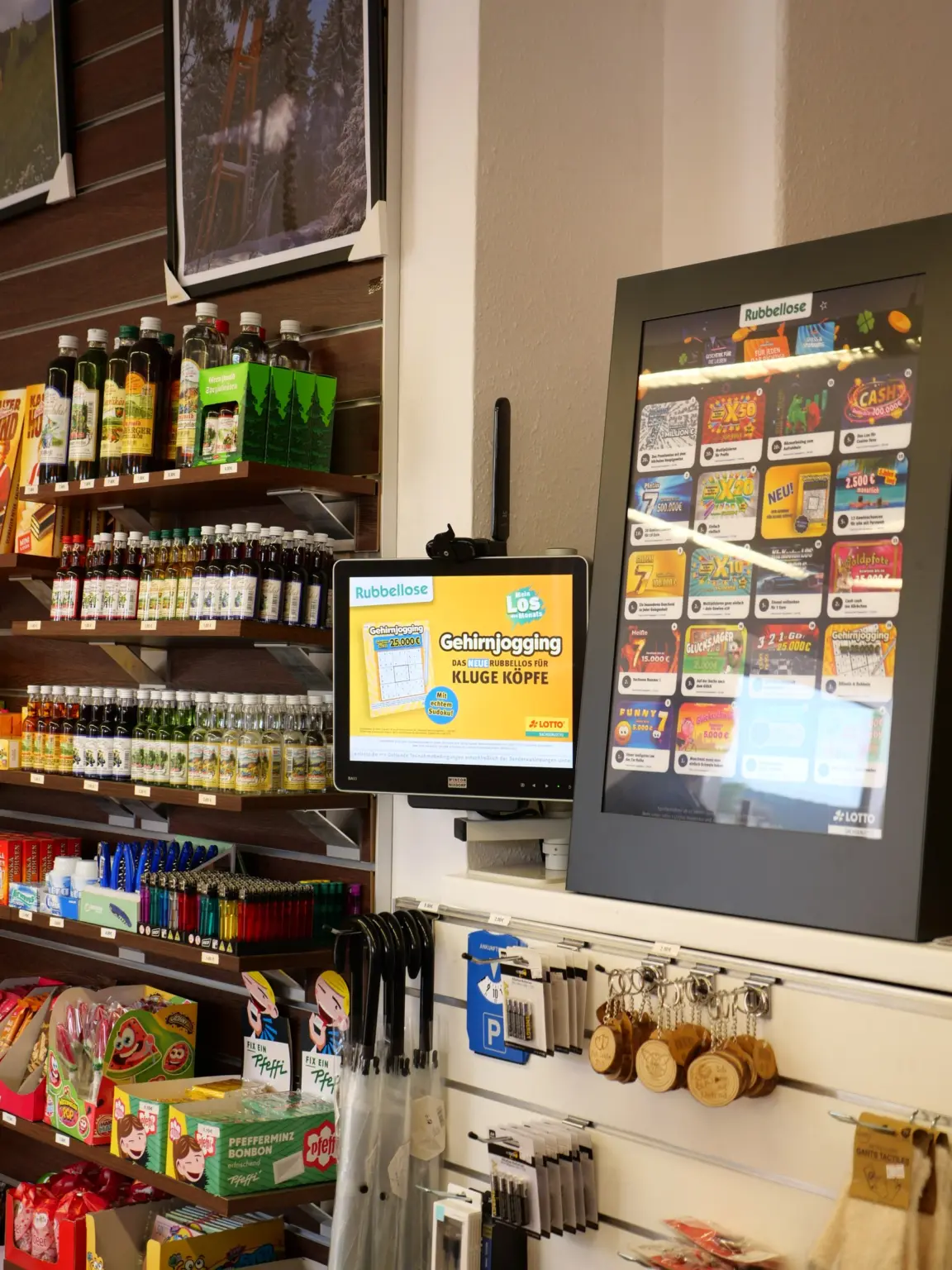 Lotto-Ecke im Laden am Markt: Bildschirm und Display mit Rubbellosen von Sachsenlotto, daneben Regale mit Kräuterlikören und Holz-Schlüsselanhängern