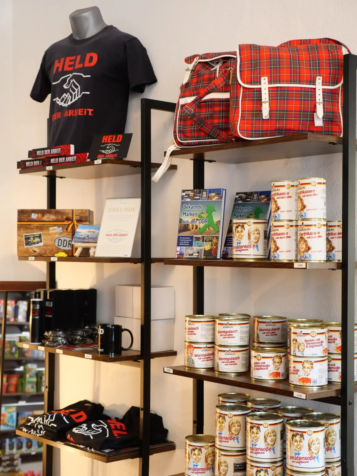 Regal mit Ostprodukten: T-Shirt „Held der Arbeit“, rot karierte Retro-Taschen, das Buch „Bekannte Marken aus der DDR“ und Konserven mit Wurstgulasch und Tomatensoße